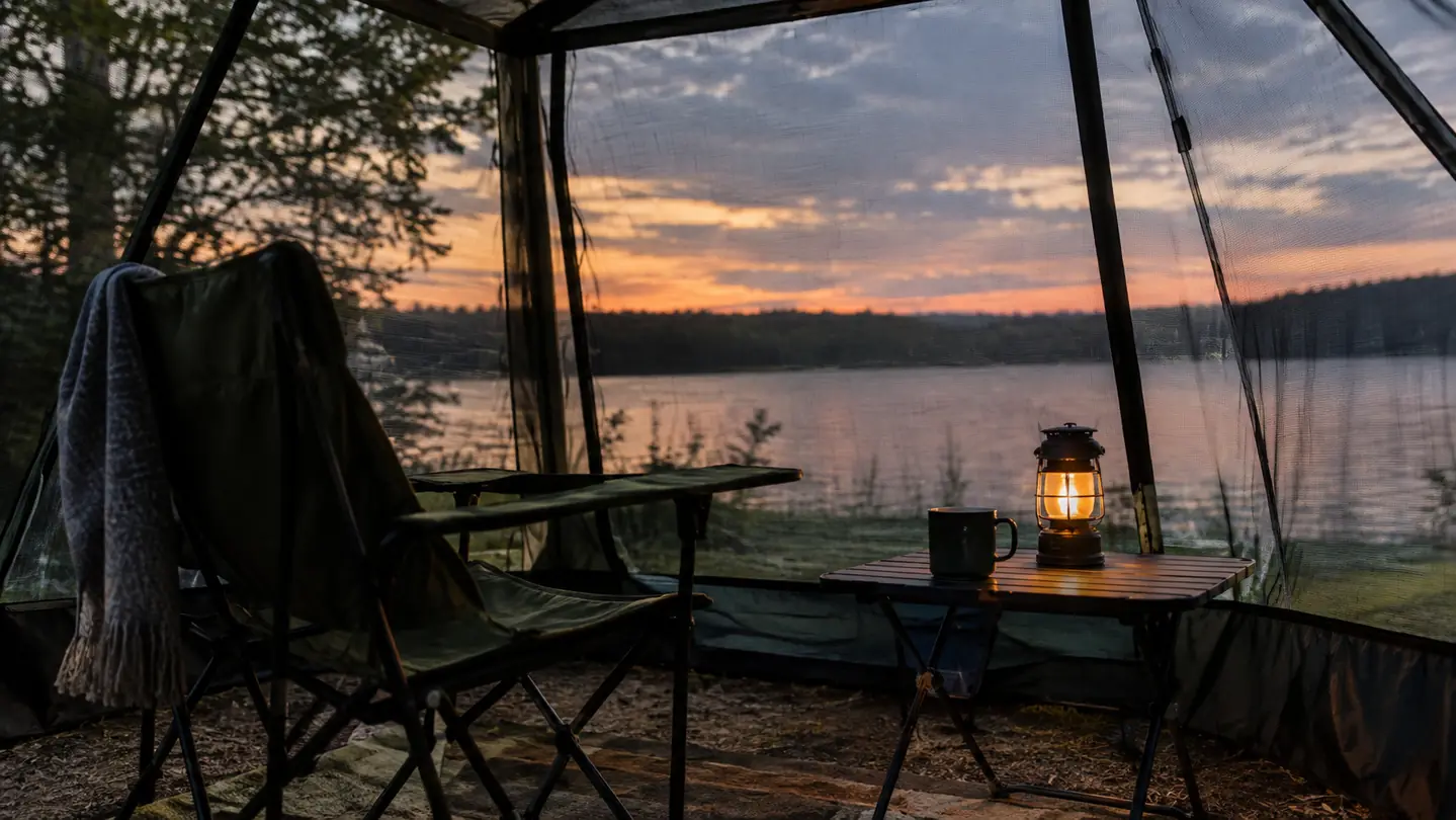 A camp chair and small table inside a screen house with a lantern and a mug